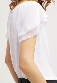 Bílá blůza s krátkými volánkovými rukávy a knoflíkovým detailem na zadní straně. Měkká, hladká tkanina, volný střih, minimalistický styl.
