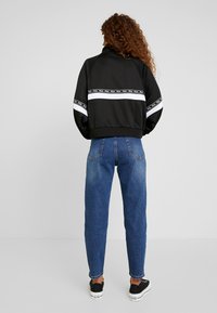 Černá bomber bunda s bílým logem, vysokým límcem a spadlými rameny, doplněná o modré džíny s vysokým pasem a černé tenisky.