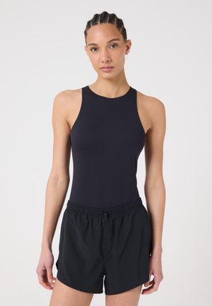 Donna con capelli intrecciati che indossa un top nero senza maniche e pantaloncini neri a vita elastica su uno sfondo chiaro uniforme.