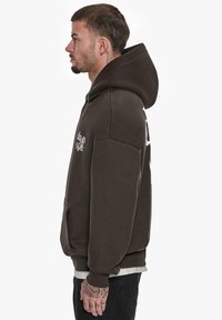 Brauner Kapuzenpullover mit einer Vorderaschlusstasche und weißen Textdetails. Verfügt über eine große Kapuze und gerippte Bündchen, mit einem entspannten Schnitt.