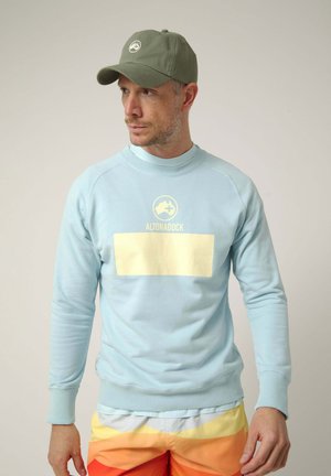Hombre con gorra verde oliva y sudadera azul claro con el logo "ALTONADOCK", combinado con pantalones cortos a rayas amarillas y naranjas, mirando hacia la izquierda.