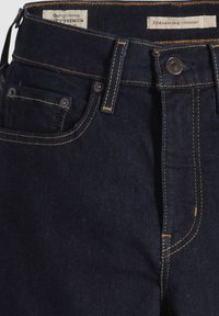 Jeans rectos de alta cintura en denim oscuro, con costuras amarillas contrastantes, diseño de cinco bolsillos y etiqueta de marca en la cinturilla.