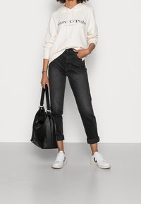Felpa chiara con logo nero, jeans neri a vita alta e sneakers bianche. Borsa nera con manici arrotolati. Abbigliamento casual.