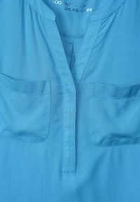 Ljusblå blus med V-hals, två bröstfickor och korta ärmar. Slätt tyg, ren sömnad och en avslappnad passform.