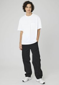 T-shirt en coton blanc avec une poche sur la poitrine, associé à un pantalon noir ample. Le mannequin porte des chaussures de sport blanches. Tenue simple et décontractée.