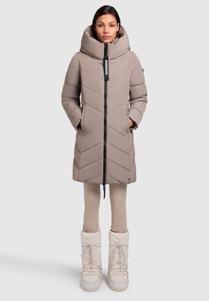 Beige pufferjas met een hoge kraag en chevron-stiksel. Voorzien van een ritssluiting en zijzakken. Draagt over lichte leggings en witte snowboots.