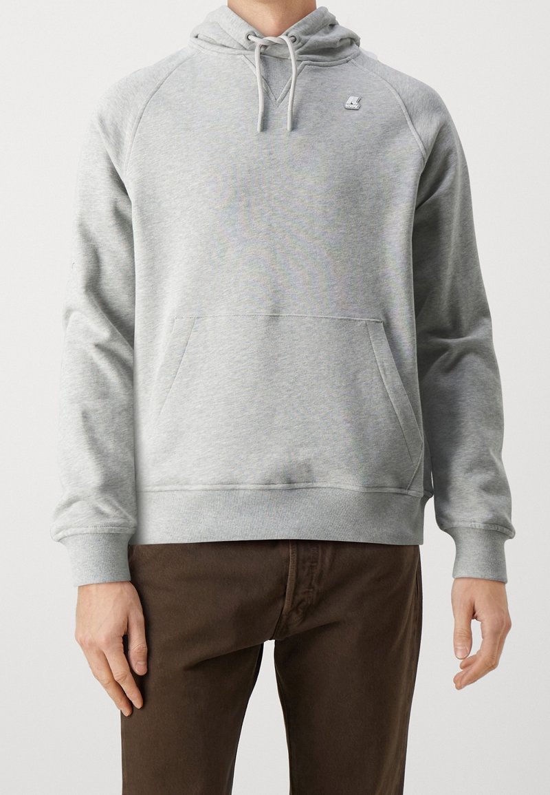 Personne portant un sweat à capuche gris clair avec une poche avant et un pantalon marron, debout devant un fond uni.
