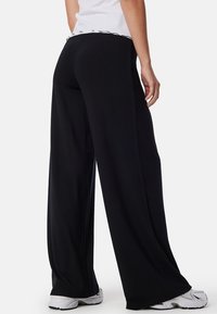 Zwarte wijde broek van zacht materiaal, met een hoge tailleband en zijnaad, gecombineerd met witte sportschoenen.