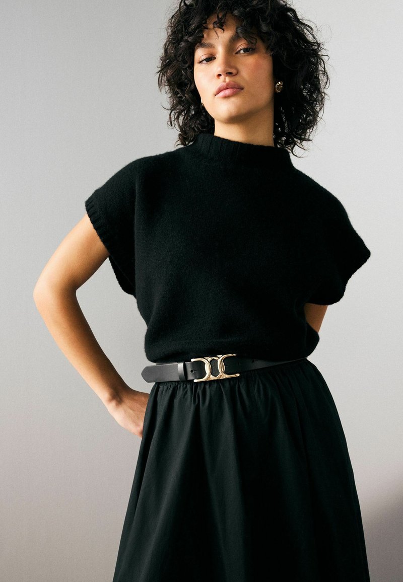 Pull noir à manches courtes avec un col haut, associé à une jupe noire. La tenue présente une ceinture avec une boucle en or.