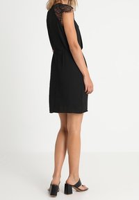 Černé šaty s krajkovými rukávy, volný střih a délka ke kolenům. Nosí se s černými sandály na blokovém podpatku. Hladká textura.