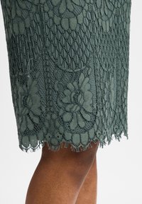 Groene kantrok met bloemenpatronen en een golvende zoom. De complexe textuur en gedetailleerde stiksels zijn prominent aanwezig.