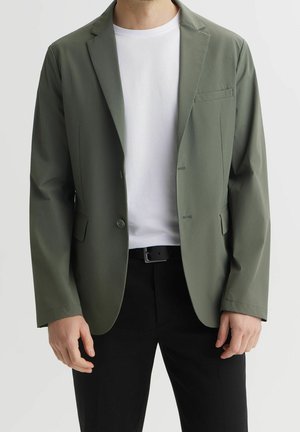 Homme portant un blazer vert olive ouvert sur un t-shirt blanc avec un pantalon noir et une ceinture noire, debout devant un fond uni.