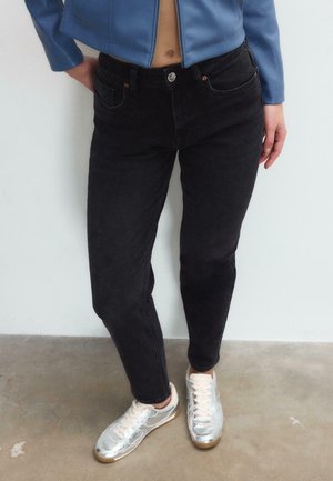 Person trägt schwarze Jeans, eine blaue kurze Jacke und silberne Turnschuhe, steht auf einem Betonboden vor einer weißen Wand.