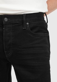Jeans de mezclilla negra con corte recto, que presentan costuras en contraste, cinco bolsillos y un botón con marca. La textura es suave con un ligero estiramiento.