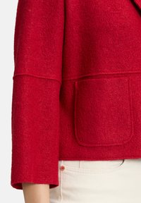 Giacca rossa in feltro con design squadrato, maniche larghe e una tasca frontale. Il tessuto testurizzato e i dettagli della semplice cucitura sono visibili.
