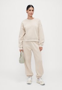 Vrouw staat in een beige sweatshirt en sweatpants, met witte sneakers, en houdt een kleine, lichtgroene handtas vast tegen een egale achtergrond.