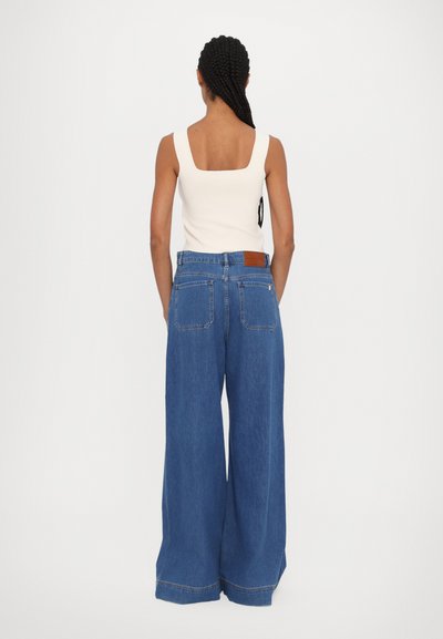 Wide-leg blue denim trousers with a high waist. Styled with a fitted, cream tank top. Features back pockets and a leather brand patch.