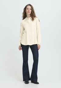 Woman with wavy hair wearing a cream long-sleeve button-up shirt, dark flared jeans, and black heeled shoes, standing against a light background.