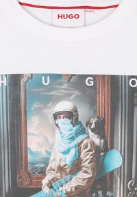 Witte t-shirt met "HUGO" label en een grafiek waarop een persoon met een helm en sjaal een snowboard vasthoudt, met een hond die achter hen zit.