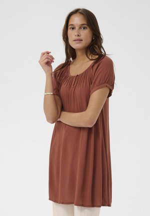 Žena s dlhými hnedými vlasmi, oblečená v voľných krátkych šatách v hrdzavej farbe, stojaca so založenou jednou rukou a druhou zdvihnutou pri tvári.