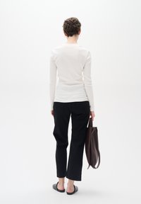 Weißes Langarm-Ripp-T-Shirt, schwarze gerade Hosen und schwarze Slipper. Hält eine braune Tragetasche. Minimalistisches Design.