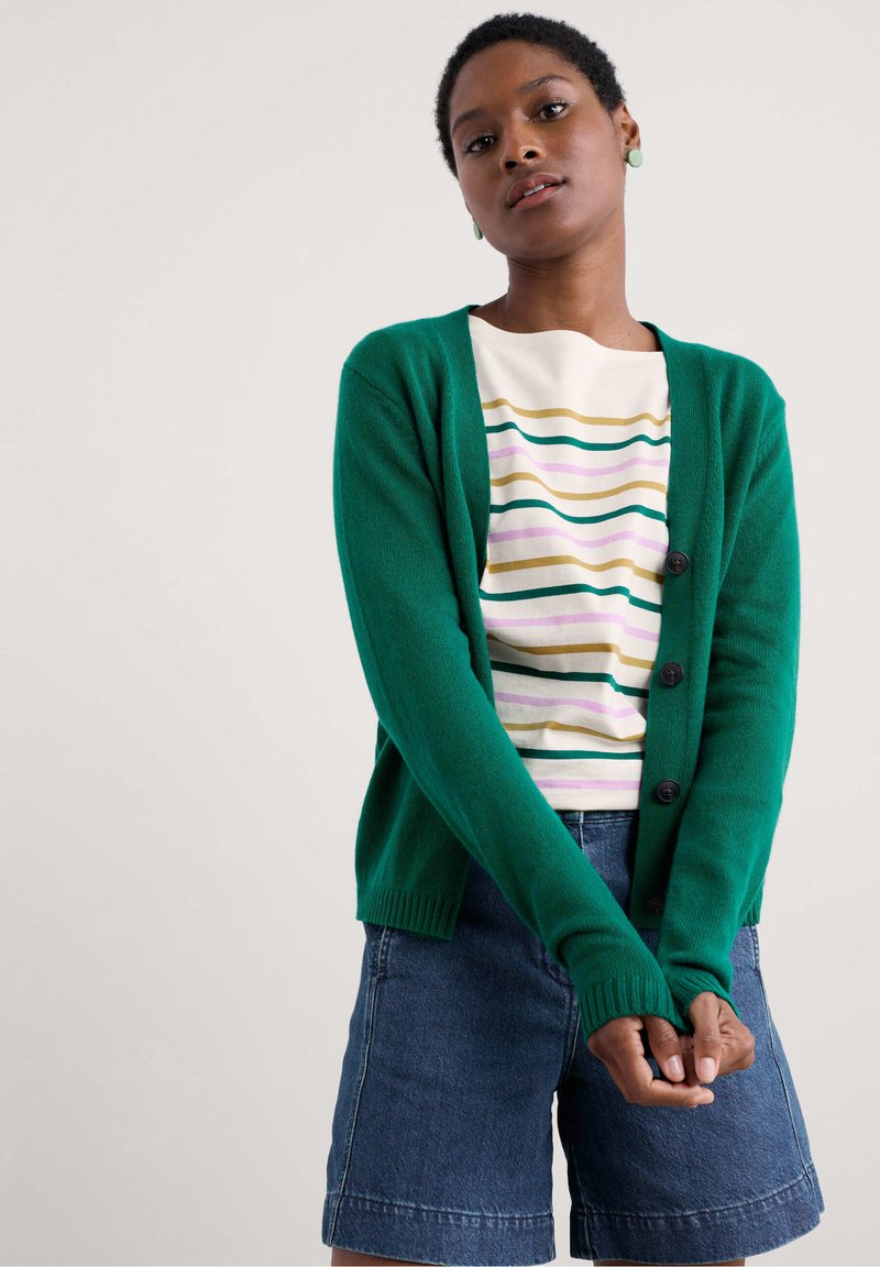 Groene gebreide cardigan met knopen aan de voorkant, geribde manchetten, gedragen over een gestreept shirt en gecombineerd met denim shorts. Eenvoudig, casual ontwerp.