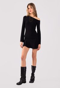 Schwarzes Langarmkleid mit schulterfreiem Design, Raffung und kurzem Rock. Kombination mit kniehohen schwarzen Stiefeln. Goldene geschichtete Kettenakzente.