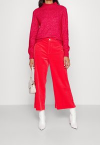 Maglione rosso a maglia con texture a coste, abbinato a pantaloni in corduroy a gamba larga di un rosso brillante. Completato con una piccola borsa color crema e stivali bianchi con tacco.