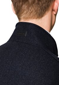 Ciemnoniebieska tkana kurtka z wysokim kołnierzem, z teksturowanym materiałem i czarną metką z tyłu blisko dekoltu.