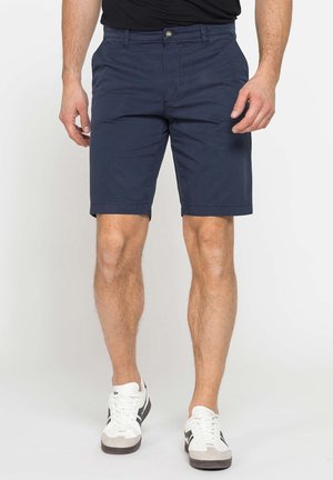 Uomo che indossa pantaloncini blu navy su misura, sneakers bianche con strisce nere e una camicia nera su uno sfondo semplice.