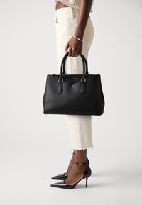 Black leather handbag with structured design, rounded handles, and gold hardware. Worn with cream trousers and pointed black heels.