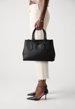 Black leather handbag with structured design, rounded handles, and gold hardware. Worn with cream trousers and pointed black heels.