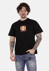 T-shirt in cotone nero con una stampa grafica colorata che presenta la parola "PROPAGANDA" in rosso. Vestibilità rilassata, maniche corte.