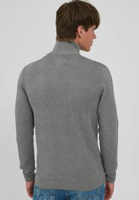 Suéter gris de punto con cuello alto, que presenta un patrón de espiga y mangas largas. La tela tiene un acabado texturizado.