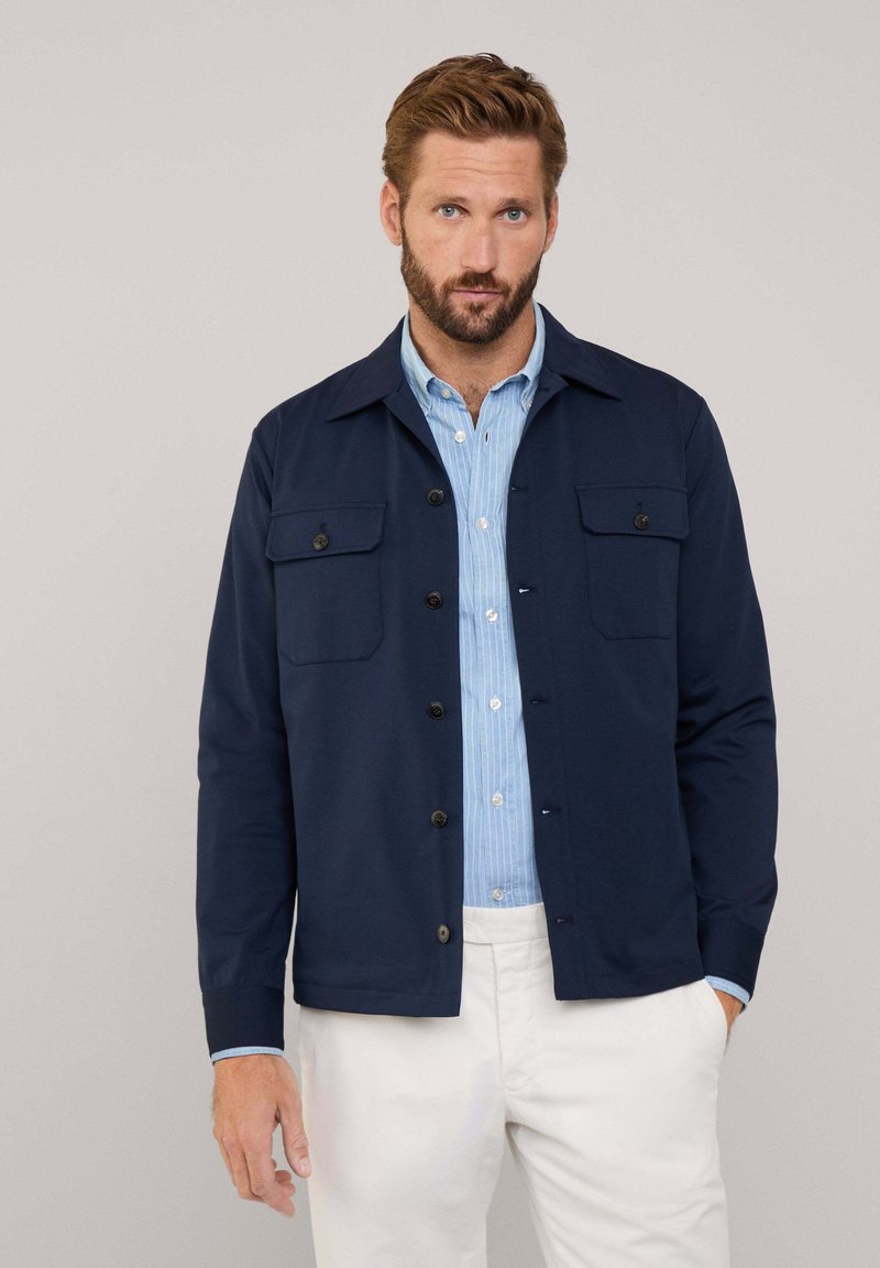 Giacca button-up blu navy realizzata in tessuto morbido, con due tasche sul petto, bottoni neri e un colletto sopra una camicia azzurra chiara.