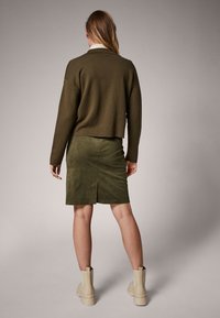 Hiais vihreä lyhyt neule yhdistettynä samanväriseen mokkahihoiseen kynnahameeseen. Mallilla on päällä beige nilkkuri, jossa on tekstuuripohja.
