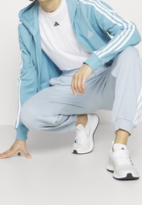 Sweat à capuche bleu avec des rayures blanches, associé à un pantalon de sport bleu clair et des chaussures de sport blanches, avec des accents noirs.