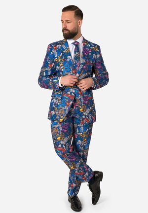 Man met baard die een blauw pak en stropdas draagt met kleurrijke robot- en voertuigpatronen, die de jasknopen aanpast en met één been over het andere staat.