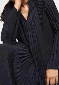 Černé plisované šaty s dlouhými rukávy, mají v-čelní výstřih a texturovanou látku, která zvýrazňuje strukturované záhyby a splývavý design.