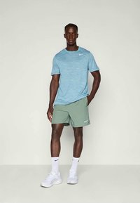 Mann steht in einem hellblauen Nike-T-Shirt, grünen Nike-Shorts, weißen Nike-Socken und weißen Sneakers vor einem schlichten Hintergrund.