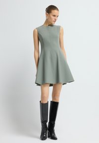 Zelené bezrukávové šaty s rozšířenou sukní, vyrobené z pevné tkaniny. V páru s černými vysokými botami s krokodýlím vzorem.