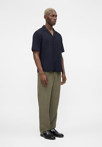 Άνδρας που στέκεται και κοιτάζει δεξιά, φοράει μαύρο πουκάμισο με κοντά μανίκια και κουμπιά, χαλαρά ελιάς πράσινα παντελόνια και μαύρα επίσημα παπούτσια.