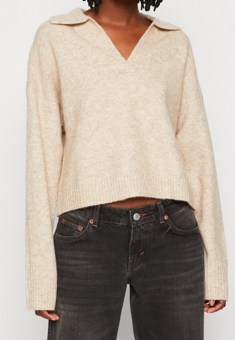 Personne portant un pull beige à col en V avec des manches longues et un jean noir taille haute, visible de la moitié de la poitrine jusqu'aux hanches.