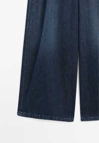 Donkerblauwe denimstof met een wijd uitlopende pijp en zichtbare stiksels langs de zoom op een effen witte achtergrond.