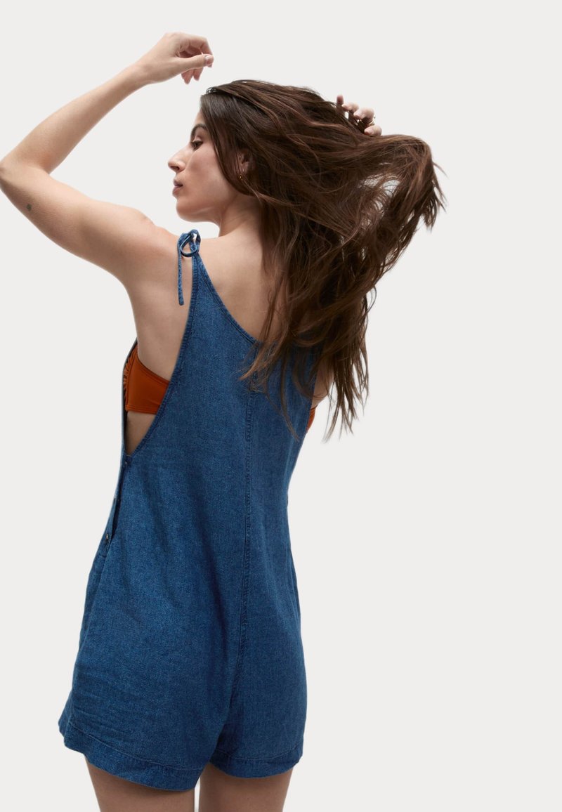 Mono de mezclilla con tirantes ajustables, lados abiertos y un corte holgado. Presenta un color azul sólido y un diseño casual.