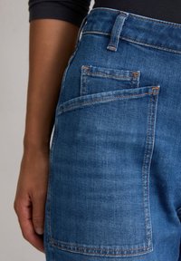 Gros plan sur une personne portant un jean en denim bleu avec une poche arrière visible et sa main reposant à côté de la hanche.