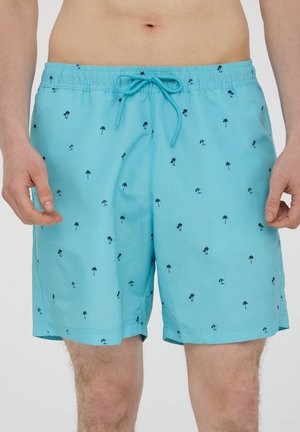 Pantaloni da bagno azzurri chiari con piccoli motivi di palme scure e ombrelloni indossati da una persona in piedi contro uno sfondo semplice.