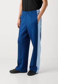 Pantalones atléticos azules con un panel lateral azul claro que presenta el texto blanco "LACOSTE". Fabricados con un tejido suave y con una cinturilla elástica.