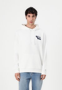 Hoodie blanc avec une poche kangourou à l'avant, arborant un logo bleu marine. Le matériau semble doux, avec des manches longues et une capuche à cordon.