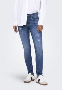 Jeans ajustados de color azul con parches desgastados, bolsillos frontales y una textura acanalada, combinados con una camiseta blanca y zapatillas blancas con detalles negros.